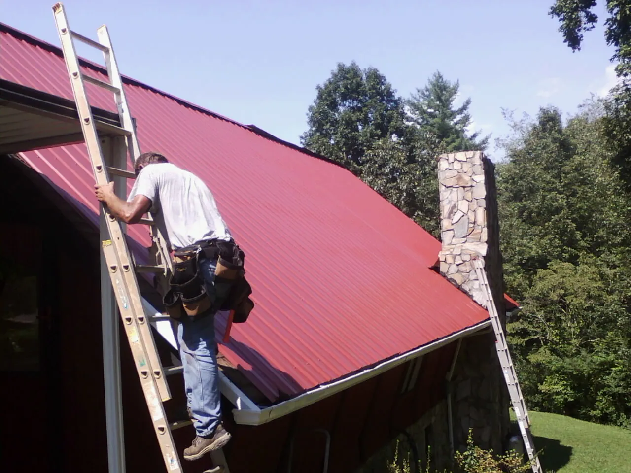Roofer on a ladder beside a red metal roof and stone chimney — Soffit & Fascia Repair by Roofing Brothers in Saugatuck